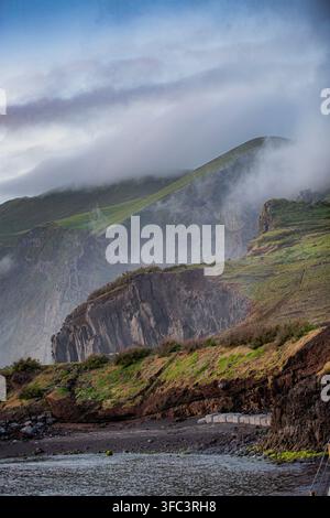 Corvo, Açores, Portugal Des falaises brumeuses s'élèvent au-dessus de pentes verdoyantes, enveloppées de brouillard. Une côte rocheuse rencontre des eaux calmes en contrebas. Les nuages tourbillonnent autour du sommet, caché Banque D'Images
