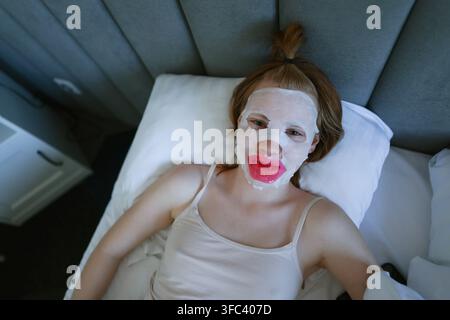 Femme allongée dans un lit portant un masque facial et un patch pour les lèvres, combinant soins de la peau, détente et mode de vie numérique. Banque D'Images