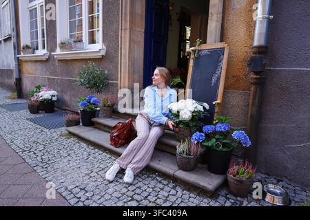 Touriste élégante assise près d'une boutique de fleurs avec des hortensias bleus et blancs dans un charmant quartier européen. Banque D'Images