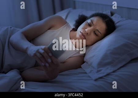 Femme noire inquiète allongée éveillée dans son lit la nuit en vérifiant son smartphone Banque D'Images