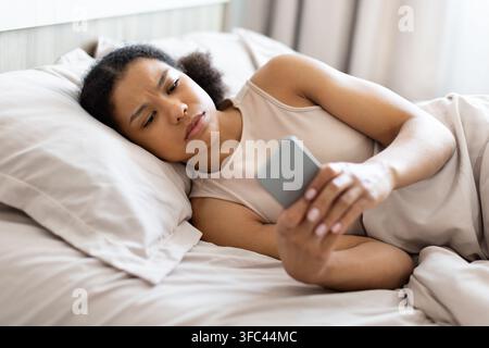 Jeune femme noire allongée dans son lit, l'air inquiète, vérifiant son smartphone Banque D'Images