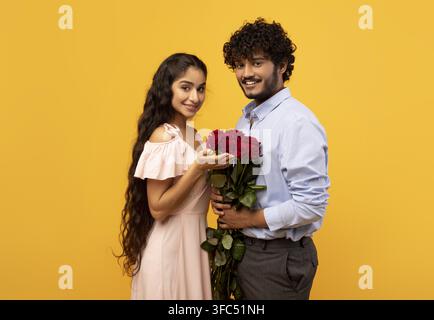 Couple sud-asiatique heureux tenant des roses, souriant et célébrant l'amour, fond jaune en studio Banque D'Images