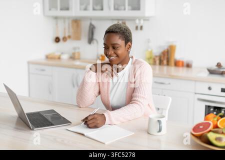 Femme noire souriante travaillant sur un ordinateur portable et prenant des notes dans une cuisine confortable, concept commercial Banque D'Images