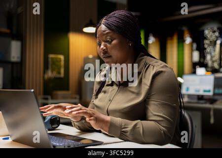 Une femme d'affaires noire participe à un appel vidéo à distance depuis son bureau sombre, maintenant un contact visuel avec la webcam. Participation à une réunion pour une conversation significative, établissant des relations et des connexions. Banque D'Images