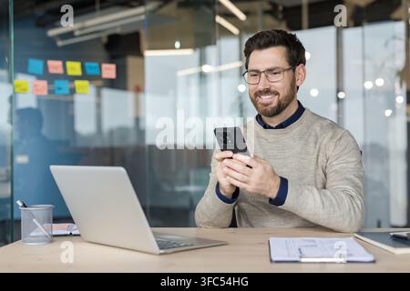 Jeune homme souriant avec barbe et lunettes assis à un bureau, regardant son téléphone portable tout en travaillant dans un environnement d'entreprise contemporain avec un ordinateur portable Banque D'Images