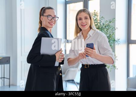 Deux femmes d'affaires souriantes avec ordinateur portable et smartphone au bureau Banque D'Images