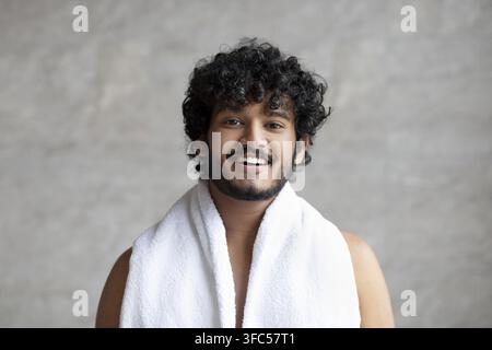 Jeune homme souriant aux cheveux bouclés portant une serviette avec une expression joyeuse, salle de bain, concept de soins de la peau Banque D'Images