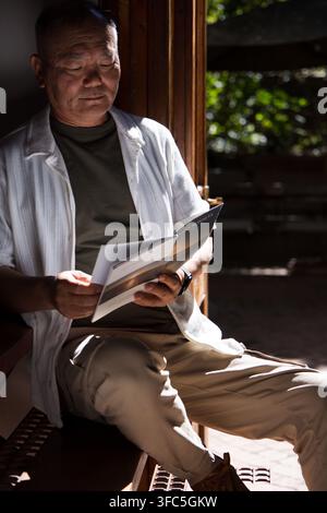Homme asiatique âgé assis sur un banc en bois près d'une porte ouverte en train de lire une brochure, la lumière du soleil sur les mains Banque D'Images