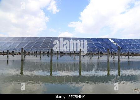 Sous un ciel bleu clair parsemé de nuages blancs, des rangées de panneaux photovoltaïques solaires sont soigneusement installées sur l'eau, formant un réseau spectaculaire. Leur réflexion Banque D'Images