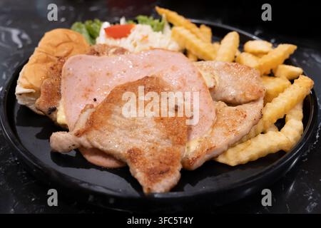 Grossissement d'un steak de porc recouvert de jambon et servi avec des frites croustillantes, du pain, de la laitue fraîche et une salade de chou crémeuse sur une assiette noire. Banque D'Images
