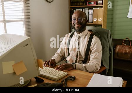 Portrait d'un homme noir souriant travaillant sur un ordinateur rétro au bureau Banque D'Images