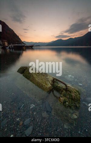Une pierre couverte de mousse avec un anneau en métal rouillé gît dans une eau claire et peu profonde d'un lac de montagne au coucher du soleil, les collines sombres se reflétant à la surface immobile. Banque D'Images
