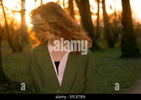Une jeune femme aux cheveux bouclés fluides vit un moment de réflexion au milieu de la lumière dorée d'un coucher de soleil dans un cadre de parc serein Banque D'Images