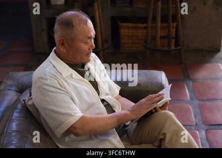 Homme asiatique âgé assis sur un canapé en cuir sombre dans un coin lecture rustique, tournant les pages d'un livre de poche Banque D'Images
