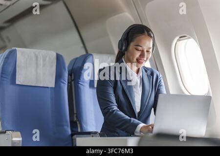 Une femme d'affaires asiatique souriante tape sur son ordinateur portable tout en portant des écouteurs dans un avion, illustrant un travail concentré et la productivité pendant les déplacements Banque D'Images