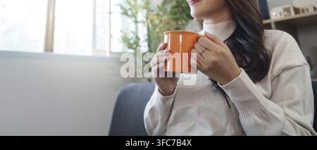 Une jeune femme tient une tasse orange et savoure une boisson chaude en se relaxant dans son salon confortable Banque D'Images