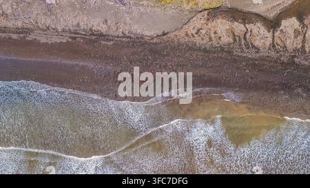 Vue aérienne des vagues de l'océan qui s'écoulent sur une plage de sable sombre couverte de déchets plastiques dispersés et de déchets en Indonésie. Banque D'Images