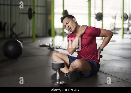 Un homme en chemise rouge est assis sur le sol de la salle de sport et se tient l'épaule inconfortablement après avoir soulevé des poids. Mise au point sur la forme physique et la prise de conscience des blessures Banque D'Images
