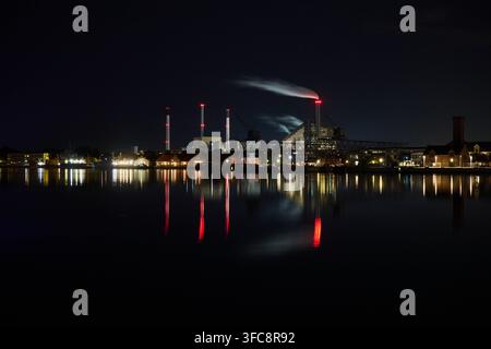 Vue nocturne de Refshaleøen à Copenhague, Danemark, avec des bâtiments industriels illuminés et de la fumée s'élevant au-dessus du front de mer. Banque D'Images