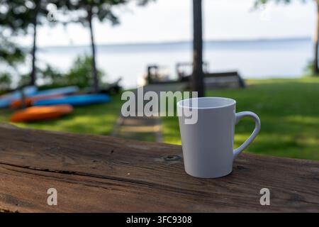 Close-up d'une tasse à café blanche sur une table en bois rustique surplombant un lac calme et une pelouse verte. Des kayaks colorés et une jetée sont doucement flous dans le b Banque D'Images