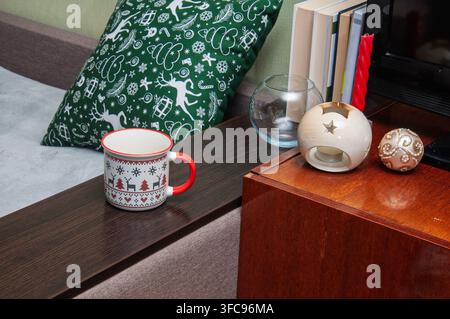 Une tasse festive repose sur une surface en bois sombre à côté d'un coussin sur le thème de Noël et d'objets décoratifs, évoquant une atmosphère chaleureuse de fêtes Banque D'Images