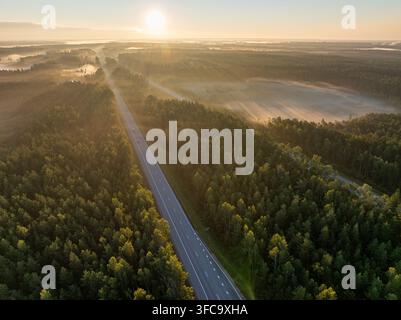 Vue aérienne de l'autoroute A2 E77 traversant un paysage forestier en Lettonie au lever du soleil avec brume matinale Banque D'Images