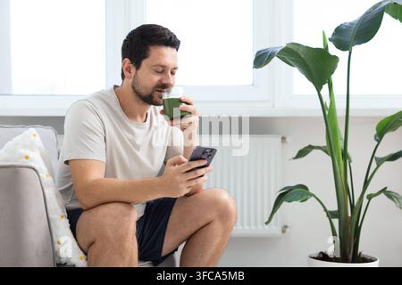 Homme avec smartphone buvant un smoothie vert dans un verre à l'intérieur Banque D'Images