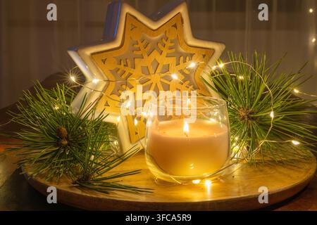 une bougie allumée dans un gobelet en verre transparent, des branches de pin fraîches, des guirlandes lumineuses et un ornement en bois en forme d'étoile de neige décoratif. Banque D'Images