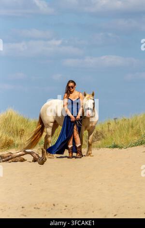 Une jeune femme en robe de soirée bleu foncé mène un cheval blanc le long d'une plage de sable sous un ciel d'été lumineux. Southport, Royaume-Uni. Banque D'Images