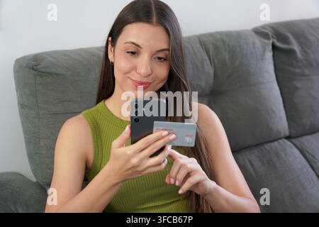 Une femme latina effectue un achat en ligne avec son smartphone et sa carte en toute tranquillité depuis son salon à la maison Banque D'Images
