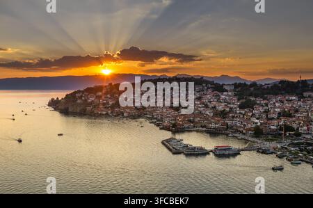 Vue aérienne de l'historique d'Ohrid au coucher du soleil, avec des maisons aux toits rouges, des bateaux dans le port et des rayons de soleil sur les montagnes à Ohrid, Municipalité d'Ohrid Banque D'Images