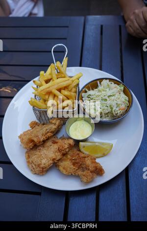 Prise verticale d'une assiette de filets de poisson frits panés croustillants servis avec des frites dorées dans un panier en métal, une salade de chou et une sauce tartare Banque D'Images