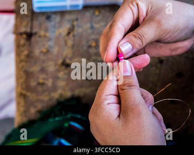 Gros plan de mains enfilant soigneusement de petites perles colorées sur un fil fin lors d'un processus traditionnel de fabrication de bijoux artisanaux. L'image met en valeur c Banque D'Images