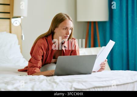 Femme travaillant sur un ordinateur portable et lisant des documents dans une chambre d'hôtel Banque D'Images