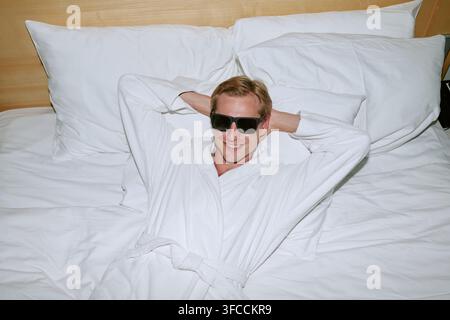 Portrait d'un homme souriant en peignoir et lunettes de soleil se détendant sur un lit d'hôtel Banque D'Images