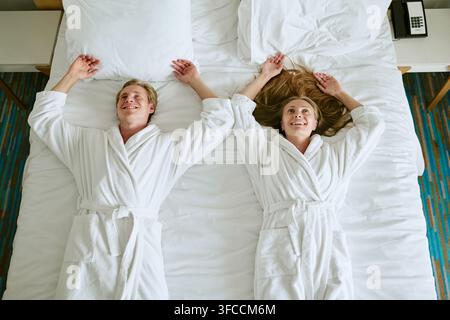 Couple heureux en peignoirs allongé sur le lit d'une chambre d'hôtel de luxe Banque D'Images