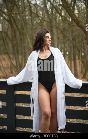 Femme en maillot de bain noir avec robe blanche sur une terrasse dans un cadre forestier. Portrait d'extérieur élégant pour les concepts de bien-être, beauté, retraite de luxe et mode de vie axée sur les soins personnels Banque D'Images
