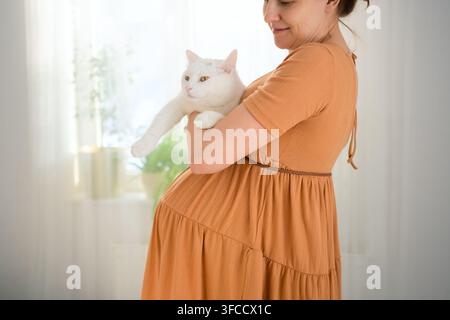Une femme enceinte tient un chat blanc dans une pièce lumineuse. La future mère enlaçant sa compagne montre une manipulation compatible et soignée ainsi qu'une compatibilité avec les animaux domestiques pendant la grossesse à la maison. Banque D'Images