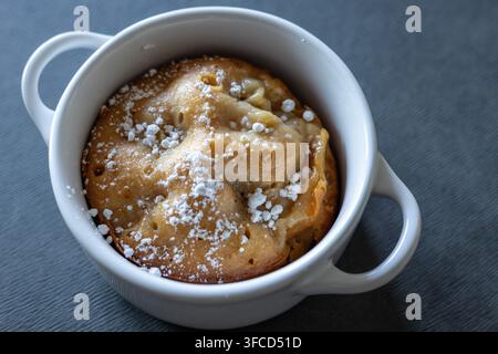 Vue inclinée d'un gâteau individuel aux pommes doré dans une cocotte blanche à deux anses, saupoudrée de sucre glace sur un tissu texturé gris Banque D'Images