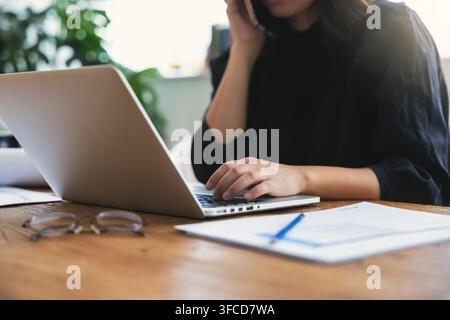 Femme d'affaires coréenne multitâchant au téléphone tout en travaillant avec un ordinateur portable à domicile Banque D'Images