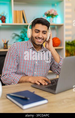 L'homme indien sur la table à la maison travaille sur ordinateur portable dans des écouteurs sans fil tapant du texte et écoutant de la musique avec une concentration calme à l'intérieur. Arabian Guy sur la table de la maison apprécie le travail freelance détendu et la routine de bureau à domicile Banque D'Images