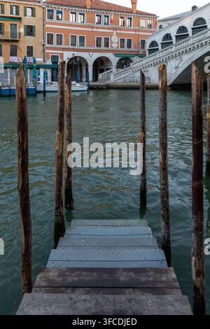 Quai en bois et poteaux d'amarrage usés à côté du pont du Rialto à Venise, Italie, avec des bâtiments historiques au bord de l'eau et une eau ondulante. Banque D'Images
