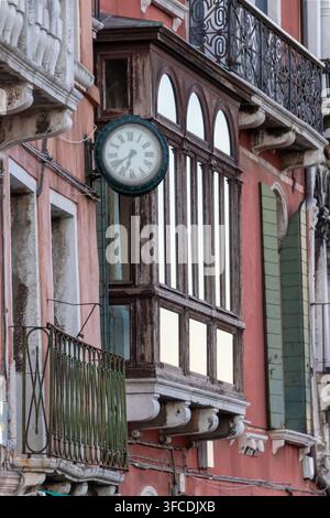Façade historique d'un bâtiment de Venise avec une horloge de rue ronde, des fenêtres en arc, des détails en bois patiné et des balcons en fer forgé en Italie. Banque D'Images