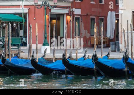 Gondoles traditionnelles amarrées le long d'un canal tranquille à Venise, Italie, avec des poteaux en bois, des bâtiments historiques rouges et une lumière chaude du soir. Banque D'Images