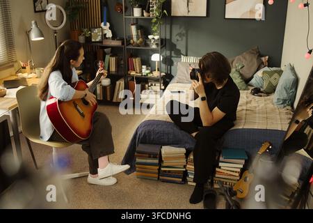 Jeune femme caucasienne jouant de la guitare acoustique tandis qu'un jeune homme asiatique prend des photos avec un appareil photo reflex dans la chambre, représentant la création de contenu et le marketing de passe-temps, prise de vue à angle élevé Banque D'Images
