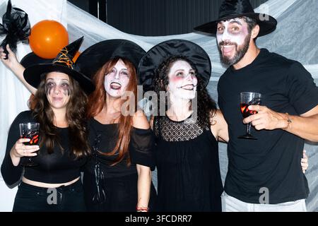 Amis adultes portant des chapeaux de sorcière et un maquillage facial effrayant, profitant de boissons et de ballons lors d'une célébration d'Halloween amusante en intérieur. Banque D'Images