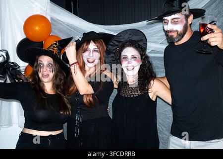Amis adultes portant des chapeaux de sorcière et un maquillage facial effrayant, profitant de boissons et de ballons lors d'une célébration d'Halloween amusante en intérieur. Banque D'Images