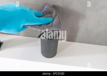 Une personne portant un gant de protection bleu essuie un distributeur de savon noir avec un chiffon en microfibres gris, mettant l'accent sur l'hygiène et la propreté Banque D'Images