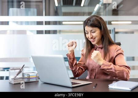 Femme d'affaires célébrant le succès à son ordinateur portable avec les poings serrés et un sourire joyeux dans un bureau moderne, montrant l'accomplissement, la motivation et la victoire professionnelle Banque D'Images