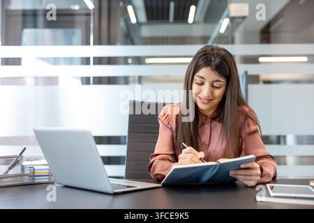Jeune femme d'affaires souriant tout en se concentrant sur l'écriture de notes dans un agenda ou un carnet, assise à un bureau équipé d'un ordinateur portable dans un environnement de bureau lumineux et contemporain Banque D'Images